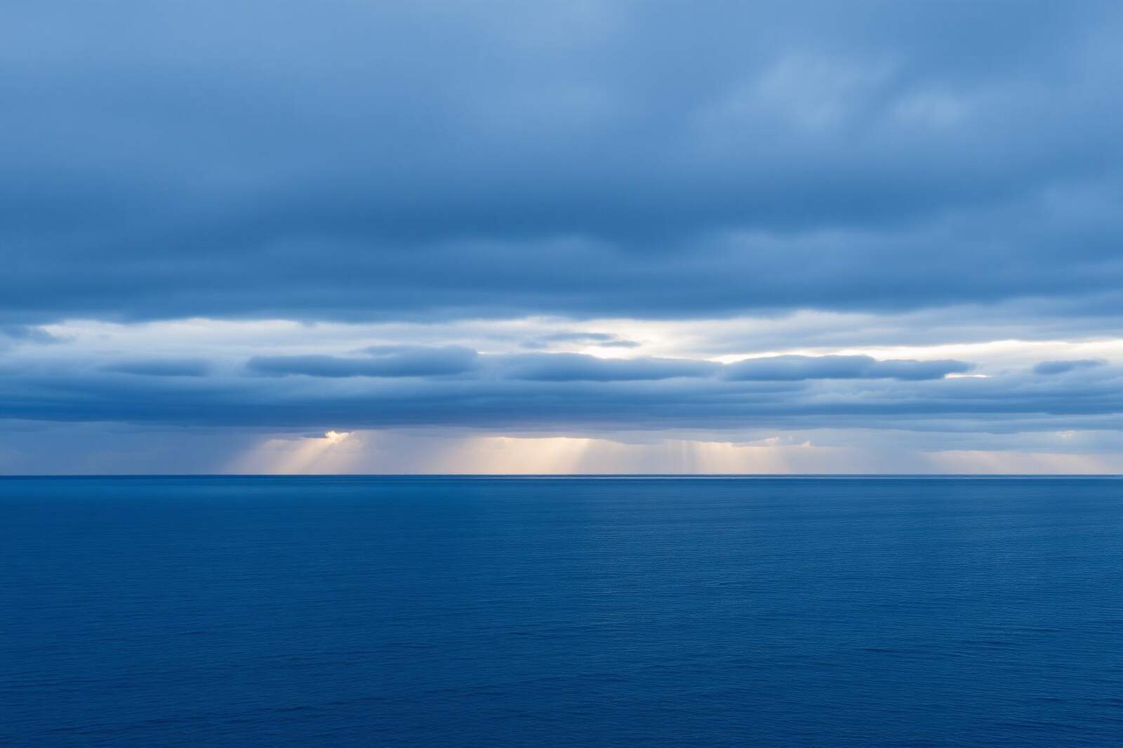 Weites, tiefblaues Meer mit geschichteten Wolken am Horizont und Lichtstrahlen zwischen den Wolken