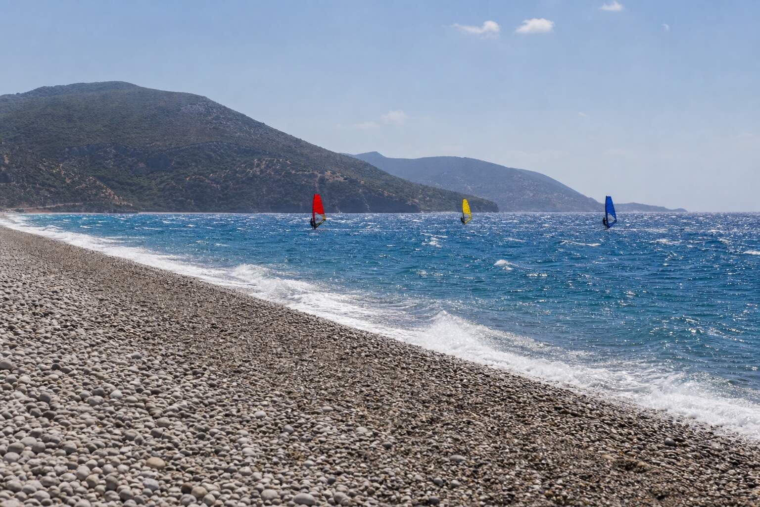 Langer Kiesstrand mit Windsurfern weit draußen auf bewegtem Meer